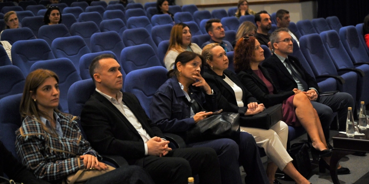 Konferans salonunda mavi koltuklarda oturan ve dikkatle bir sunumu izleyen katılımcı grubu. Ön sırada, şık kıyafetler giymiş kadın ve erkeklerden oluşan bir grup yer alıyor; bazıları ellerinde telefon tutuyor veya not alıyor. Salonun geri kalanında da izleyiciler bulunuyor.