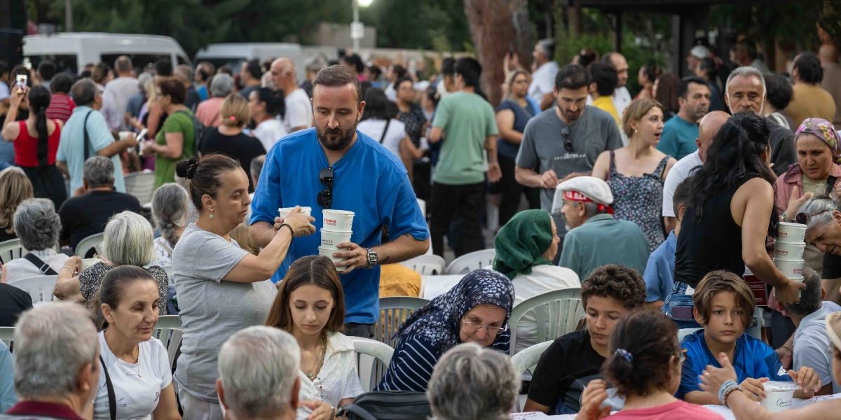 Açık havada düzenlenen bir toplu yemek etkinliğinde, mavi tişörtlü bir adam elinde üst üste dizili Bornova Belediyesi logolu yemek kaplarını masalarda oturanlara dağıtıyor. Beyaz plastik sandalyelerde oturan farklı yaş gruplarından çok sayıda insan, önlerindeki masalarda yemeklerini yiyor. Arka planda ayakta duran ve sohbet eden kalabalık bir grup daha bulunuyor.