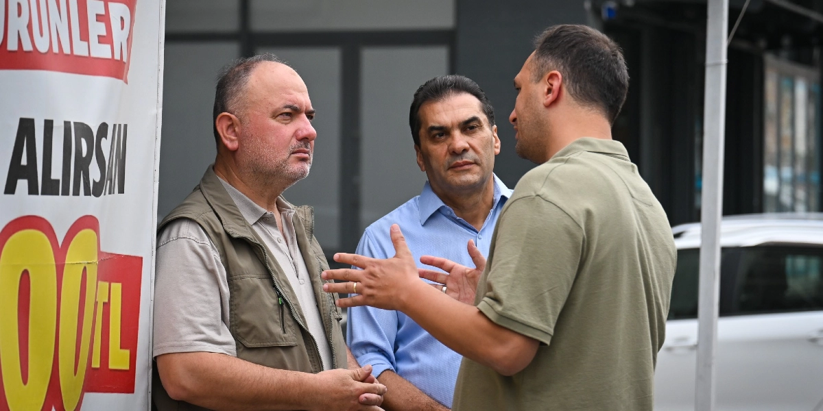 Haki renk yelekli bir adam, açık mavi gömlekli bir adam ve haki renk polo yaka tişörtlü bir adam sokakta bir arada durarak sohbet ediyor. Sol tarafta üzerinde büyük harflerle 