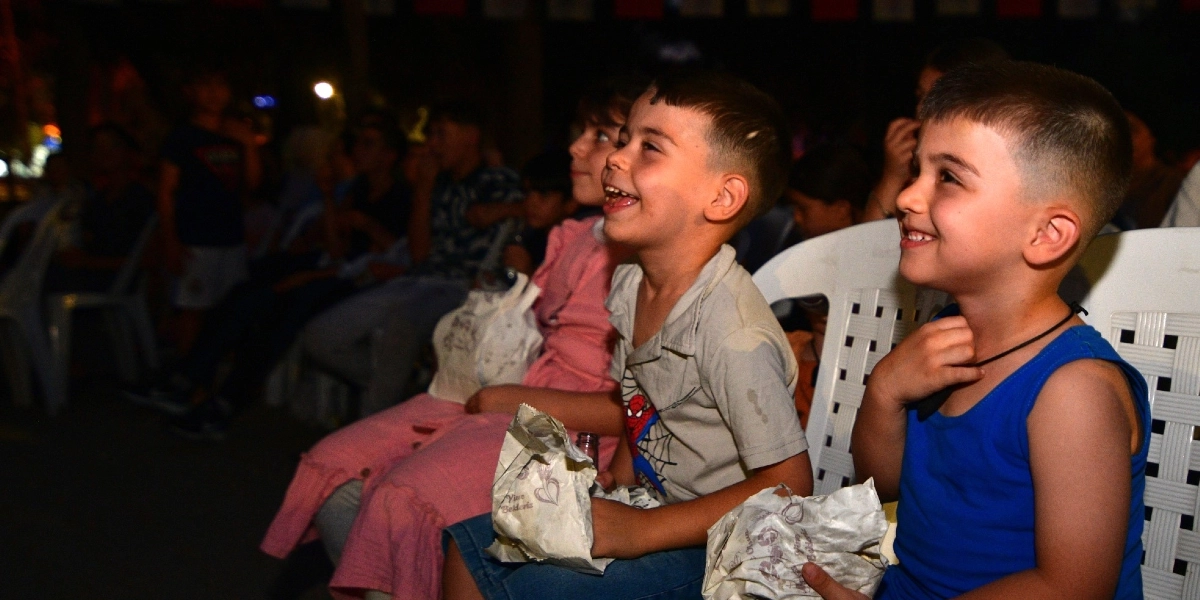Açık havada düzenlenen bir sinema etkinliğinde, beyaz plastik sandalyelerde yan yana oturan ve neşeyle ekrana bakan çocuklar. Ön planda, üzerinde Örümcek Adam baskılı tişört olan ve ağzı açık gülen bir çocuk ile yanında mavi atlet giymiş, gülümseyen bir başka çocuk yer alıyor. Çocukların kucaklarında kağıt paketler bulunuyor ve arkalarında etkinliği izleyen diğer izleyiciler seçiliyor.