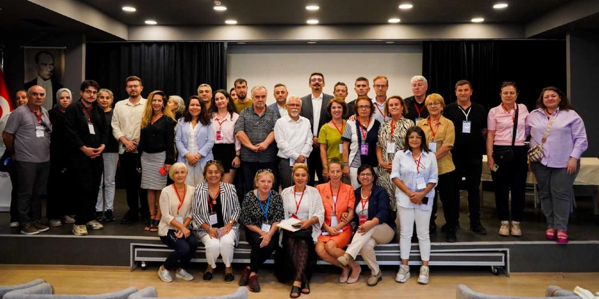 Kapalı bir salonda, sahne üzerinde yan yana dizilmiş, farklı yaş ve giyim tarzlarına sahip yaklaşık 30 kişilik bir grup, fotoğraf çekimi için kameraya gülümseyerek bakıyor. Grubun ön sırasında oturanlar ve arkasında ayakta duranlar bulunuyor. Katılımcıların birçoğunun boynunda etkinlik kartı asılı. Sahnenin sol tarafında duvarda Mustafa Kemal Atatürk'ün portresi ve Türk bayrağı yer alıyor. Arka planda siyah perdeler ve tavanda spot aydınlatmalar bulunuyor.