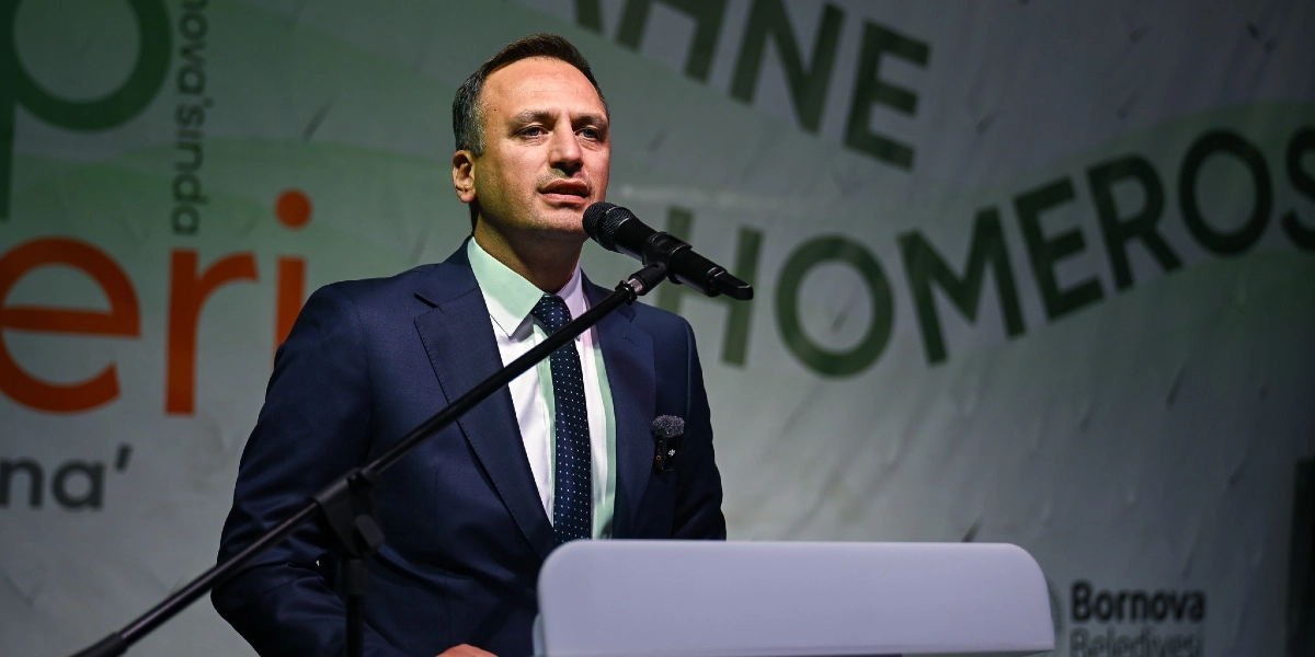 Lacivert takım elbiseli ve puantiyeli kravatlı bir erkek, arkasında Bornova Belediyesi logosunun ve Homeros temalı yazıların yer aldığı bir sahnede, kürsüde mikrofon başında konuşma yapıyor.