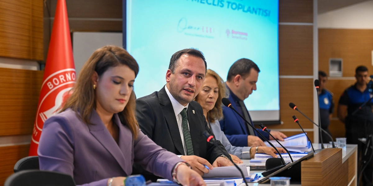 Bornova Belediye Başkanı Ömer Eşki ve belediye meclis üyeleri, üzerinde Bornova Belediyesi logosunun bulunduğu bir toplantı salonunda, önlerinde belgelerle bir meclis toplantısında oturuyorlar. Arka planda 