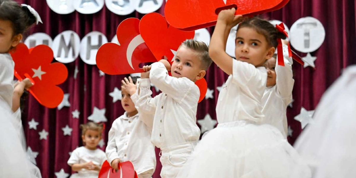 Beyaz kıyafetler giymiş anaokulu öğrencileri, ellerinde üzerinde Türk bayrağı figürü bulunan kırmızı kalp şeklinde panolarla sahnede gösteri yapıyor. Çocukların arkasında bordo renkli bir perde ve üzerinde beyaz yıldızlar ile harflerden oluşan bir süsleme yer alıyor.