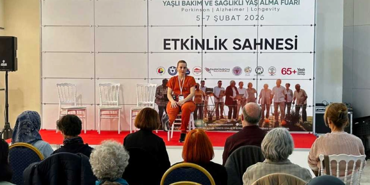 Yaşlı Bakım ve Sağlıklı Yaş Alma Fuarı kapsamında düzenlenen etkinlik sahnesinde, turuncu kıyafetli bir konuşmacı sandalyede oturarak izleyicilere hitap ediyor. Sahne arkasındaki büyük panoda 5-7 Şubat 2026 tarihleri, etkinlik başlığı ve çeşitli kurum logoları yer alıyor. İzleyiciler, sahne önündeki sandalyelerde oturarak konuşmacıyı dinliyor.