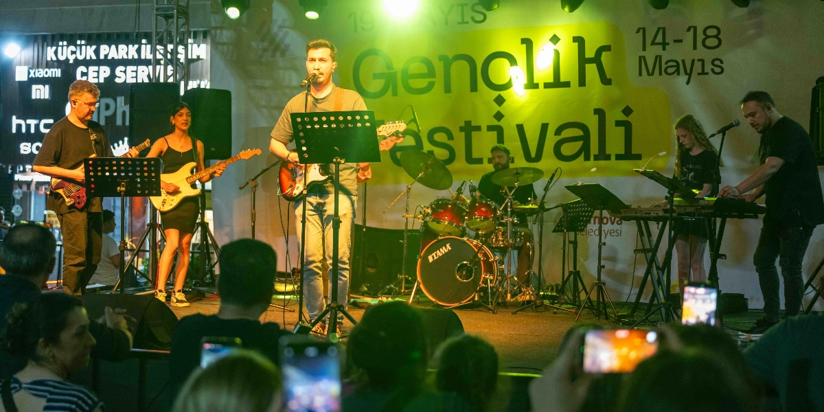Sahne üzerinde canlı performans sergileyen beş kişilik bir müzik grubu; solda elektro gitar çalan bir erkek ve bir kadın, ortada mikrofon başında şarkı söyleyen ve elektro gitar çalan bir erkek, arkada bateri çalan bir davulcu, sağda ise klavye çalan bir kadın ve bir erkek müzisyen yer alıyor. Arka planda 