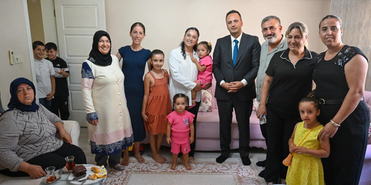 Ev ortamında bir araya gelmiş, aralarında kadınların, çocukların ve takım elbiseli bir erkeğin bulunduğu kalabalık bir grup. Sol tarafta oturan başörtülü bir kadın, önündeki sehpada bulunan çay ve tatlı tabağıyla birlikte yer alıyor. Arka planda ayakta duran grup içerisinde, koyu renk takım elbiseli ve mavi kravatlı bir erkek, diğer yetişkinler ve çocuklar ile birlikte gülümsüyor. Çocuklardan biri kucağında taşınırken, diğerleri grubun önünde veya yanında duruyor. Evin iç mekanında, açık renkli bir koltuk ve desenli bir halı üzerinde gerçekleşen bu buluşmada herkes kameraya bakıyor.
