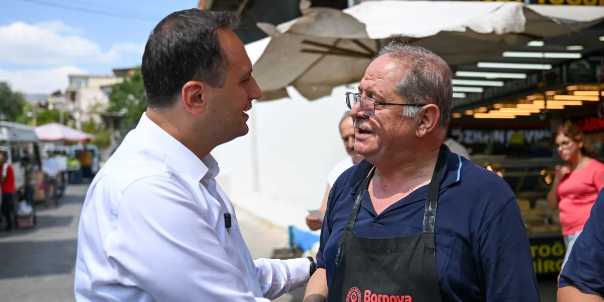 Beyaz gömlekli bir yetkili, açık hava pazarında üzerinde Bornova Belediyesi logolu siyah bir önlük bulunan, gözlüklü ve bıyıklı bir esnafla samimi bir şekilde sohbet ediyor.