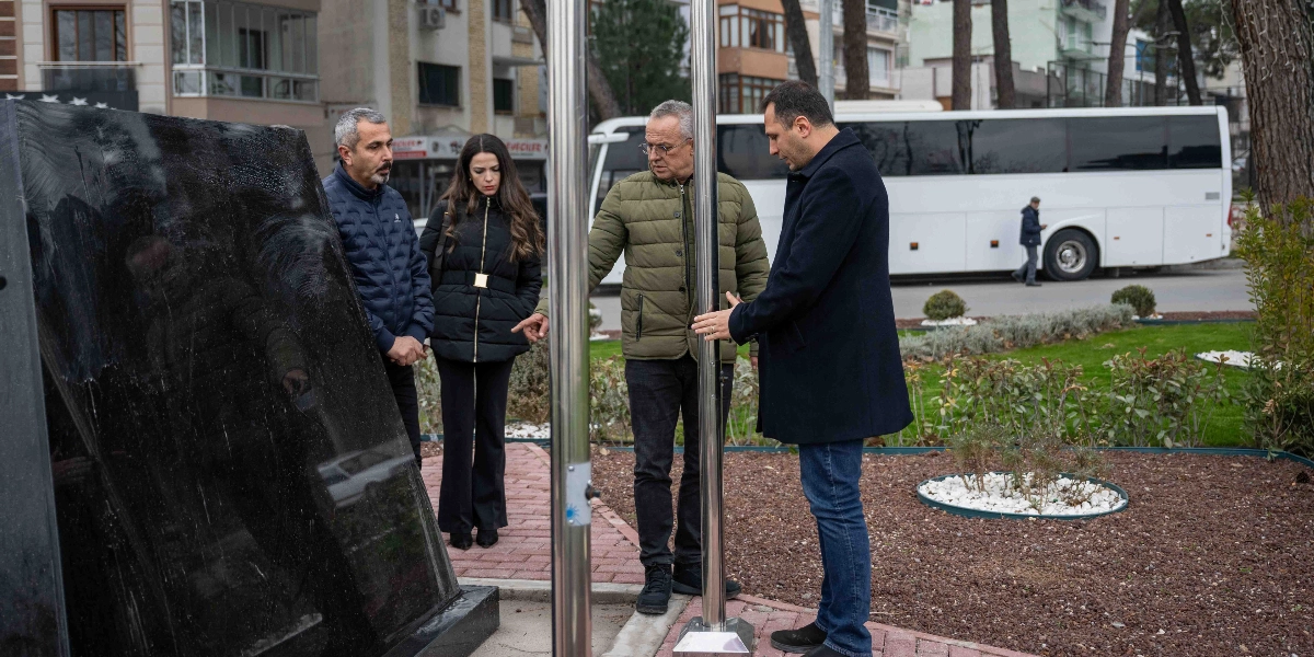 Dış mekanda, park benzeri bir alanda bulunan dört kişilik bir grup, siyah mermer bir anıtın önünde durarak incelemelerde bulunuyor. Grupta lacivert şişme montlu bir erkek, siyah kabanlı bir kadın, haki renkli şişme montlu bir erkek ve koyu renkli bir palto giymiş başka bir erkek yer alıyor. Grup üyeleri, metal bir direğin ve anıtın bulunduğu bölgeyi dikkatle incelerken, arka planda beyaz bir otobüs ve ağaçlıklı bir yol görülüyor.
