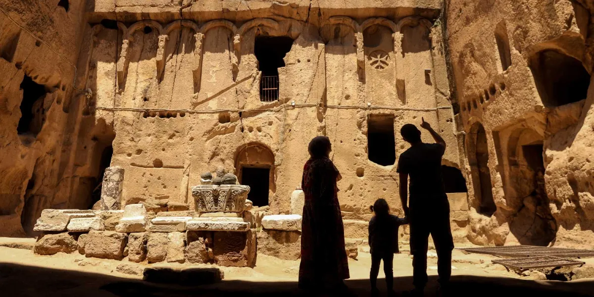 Kayalara oyulmuş antik bir yapının önünde duran bir aile, tarihi duvarları ve üzerindeki kabartmaları inceliyor. Silüet halindeki yetişkinler ve bir çocuk, mağara benzeri yapının içindeki oyuklara ve duvardaki dairesel bir haç motifine bakıyor. Ön planda ise antik dönemden kalma taş sütun başlıkları ve mimari kalıntılar yer alıyor.