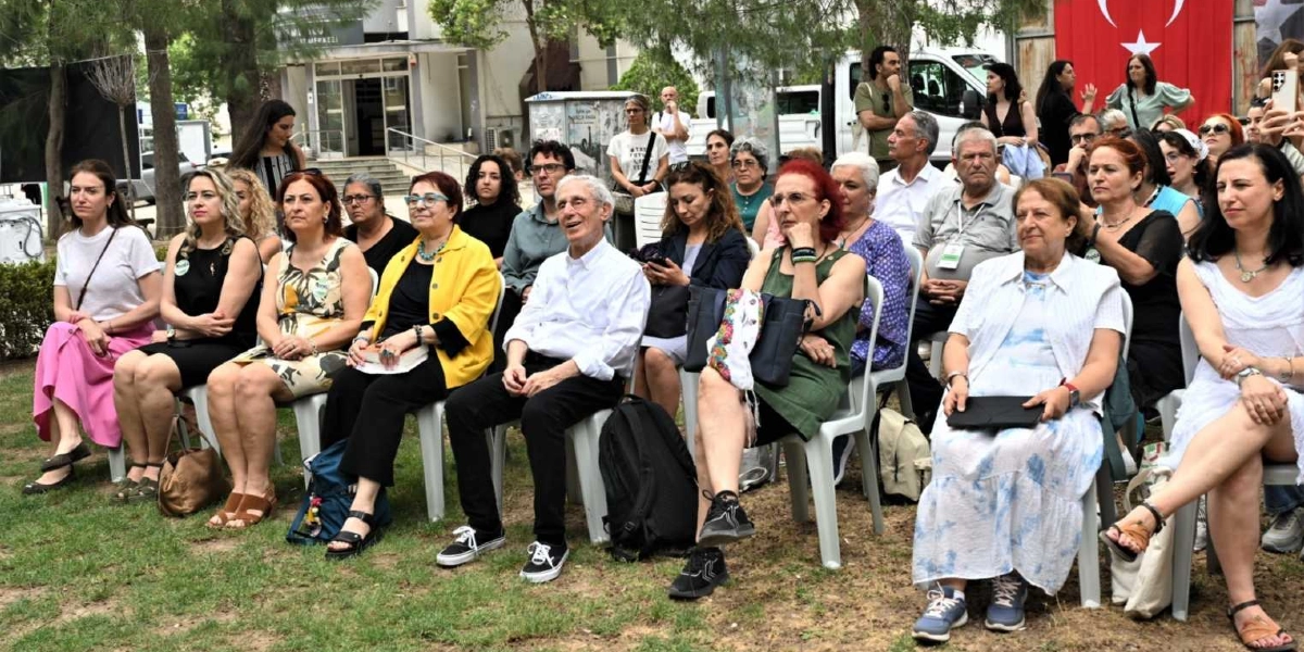 Açık havada, çimenlik bir alanda plastik sandalyelerde yan yana oturmuş, bir etkinliği dikkatle izleyen çoğunluğu kadınlardan oluşan kalabalık bir grup. İzleyiciler arasında beyaz gömlekli yaşlı bir adam, sarı ceketli bir kadın ve beyaz elbiseli kadınlar dikkat çekiyor. Arka planda ağaçlar, bir bina girişi ve asılı duran büyük bir Türk bayrağı görülüyor.