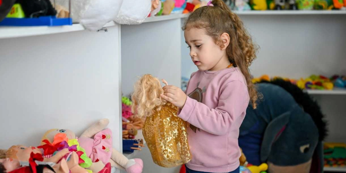 Kıvırcık saçlı küçük bir kız çocuğu, oyuncaklarla dolu bir rafın önünde elinde tuttuğu altın rengi parıltılı elbiseli bir oyuncak bebeği dikkatle inceliyor.