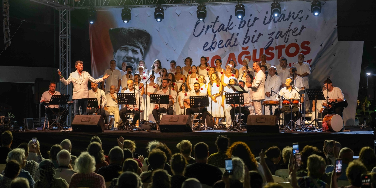 Açık havada kurulan bir sahnede, beyaz kıyafetler giymiş bir koro ve orkestra 30 Ağustos Zafer Bayramı kutlaması kapsamında konser veriyor. Sahnenin arka planında dev bir Atatürk portresi ve üzerinde 