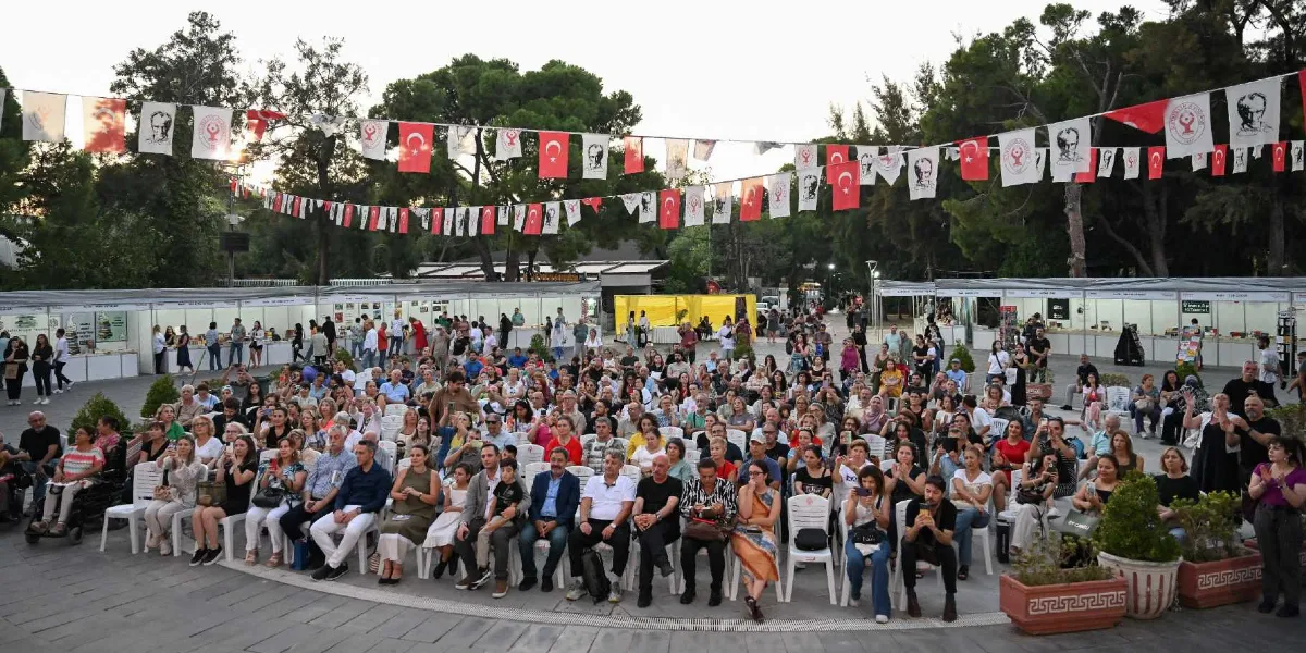 Açık havada düzenlenen bir etkinlik alanında, beyaz plastik sandalyelerde oturan kalabalık bir izleyici grubu görülüyor. Etkinlik alanı, üzerinde Türk bayrakları ve Atatürk görselleri bulunan flamalarla süslenmiş. Arka planda, kitap stantlarının yer aldığı beyaz çadırlar ve ağaçlıklı bir park alanı bulunuyor. İzleyiciler dikkatle karşıya bakarken, bazıları ellerindeki telefonlarla fotoğraf çekiyor.
