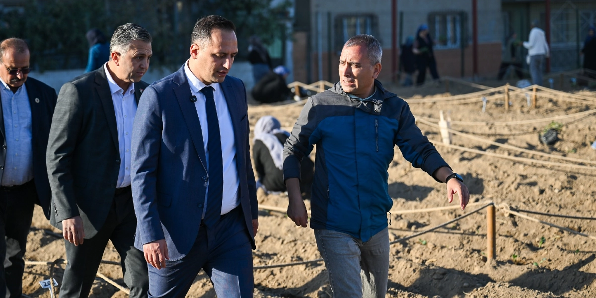 Üç erkek, toprakla çevrili bir alanda yürürken bir şeyler konuşuyor. Öndeki iki kişi lacivert takım elbiseli, arkalarındaki kişi ise gri ceketli ve gözlüklüdür. En sağdaki, mavi-gri spor bir ceket giymiş olan kişi, eliyle alanı işaret ederek açıklama yapıyor. Arka planda ise toprak zeminde iplerle ayrılmış bölümlerde çalışan ve bitki diken insanlar görülüyor.