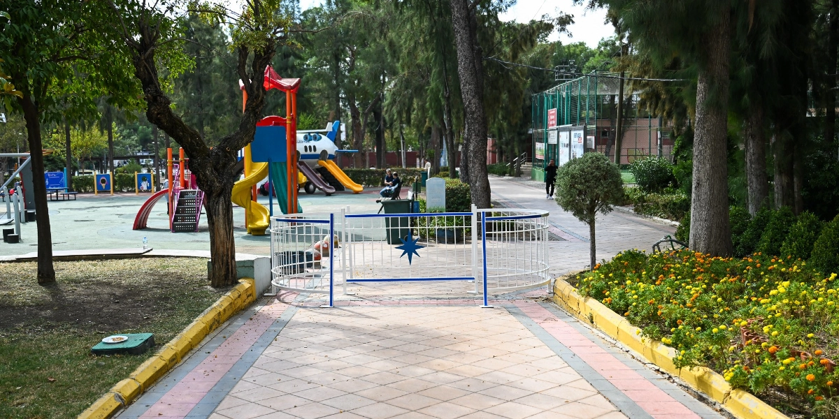 Bornova'daki bir parkın girişinde, motosikletlerin geçişini engellemek amacıyla yaya yolunun ortasına yerleştirilmiş beyaz renkli, metal bir bariyer bulunuyor. Bariyerin arkasında çocuk oyun alanı, uçak şeklinde bir kaydırak, ağaçlar ve çiçek tarhları yer alıyor.