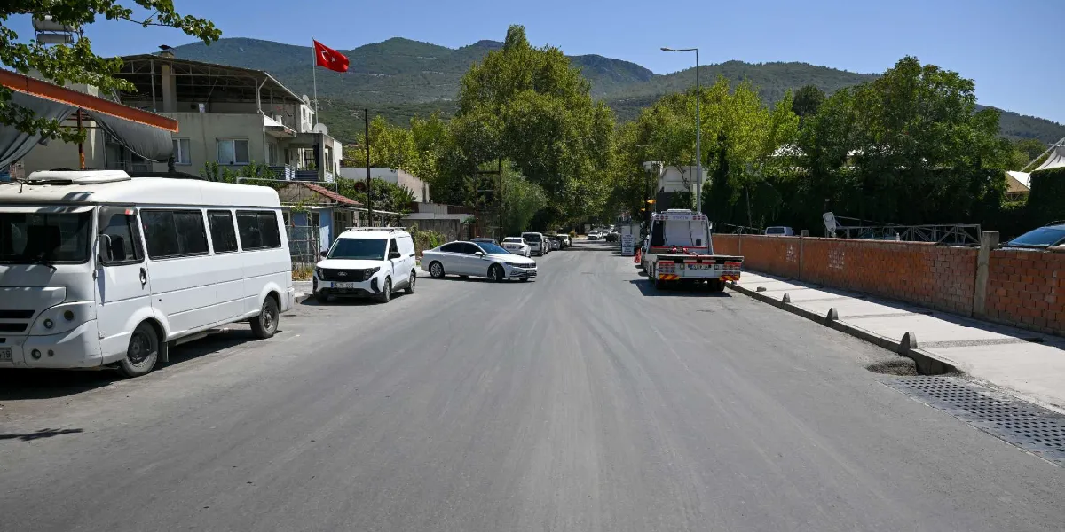 Asfaltı yeni dökülmüş, geniş ve boş Pınar Caddesi'nin iki yanında park halindeki araçlar, ağaçlar ve arka planda dağ manzarası görülüyor. Yolun sağ tarafında tuğla bir duvar ve yeni düzenlenmiş bir kaldırım bulunurken, yolun solunda ise beyaz bir minibüs ve binalar yer alıyor.