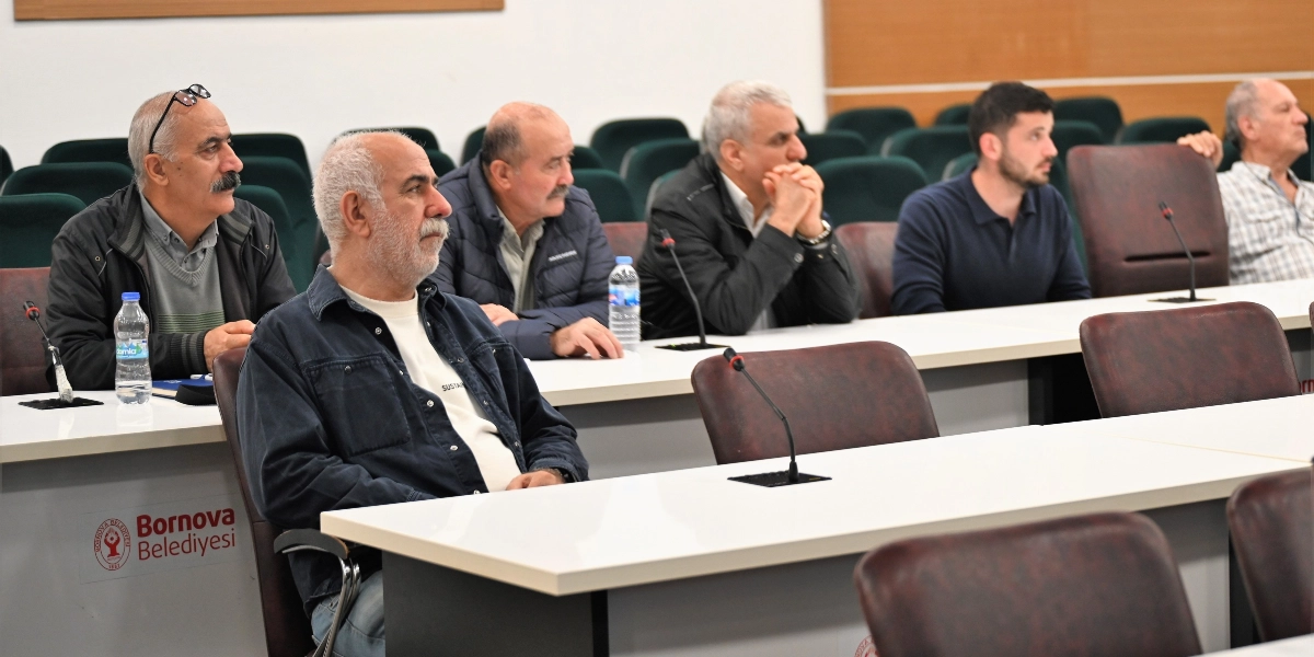 Bornova Belediyesi'ne ait bir toplantı salonunda, beyaz uzun bir masanın etrafında yan yana oturan altı erkek katılımcı dikkatle karşıya bakıyor. Katılımcıların önünde mikrofonlar ve su şişeleri bulunuyor; masanın yan tarafında ise Bornova Belediyesi logosu yer alıyor.