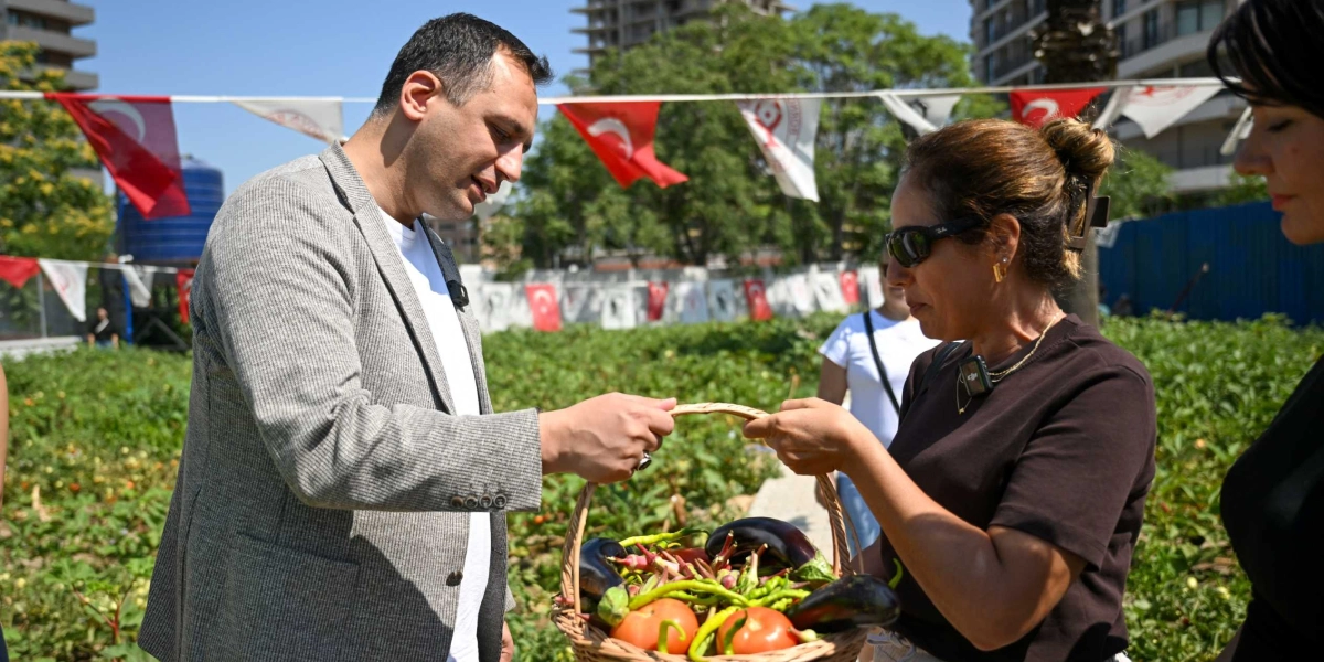 Açık havada, yeşil bir sebze bahçesinde, gri ceketli bir erkek ve kahverengi tişörtlü bir kadın, içinde patlıcan, domates ve biber bulunan hasır bir sepeti birlikte tutuyor. Arka planda Türk bayrakları asılı ve uzakta yüksek apartmanlar görülüyor.