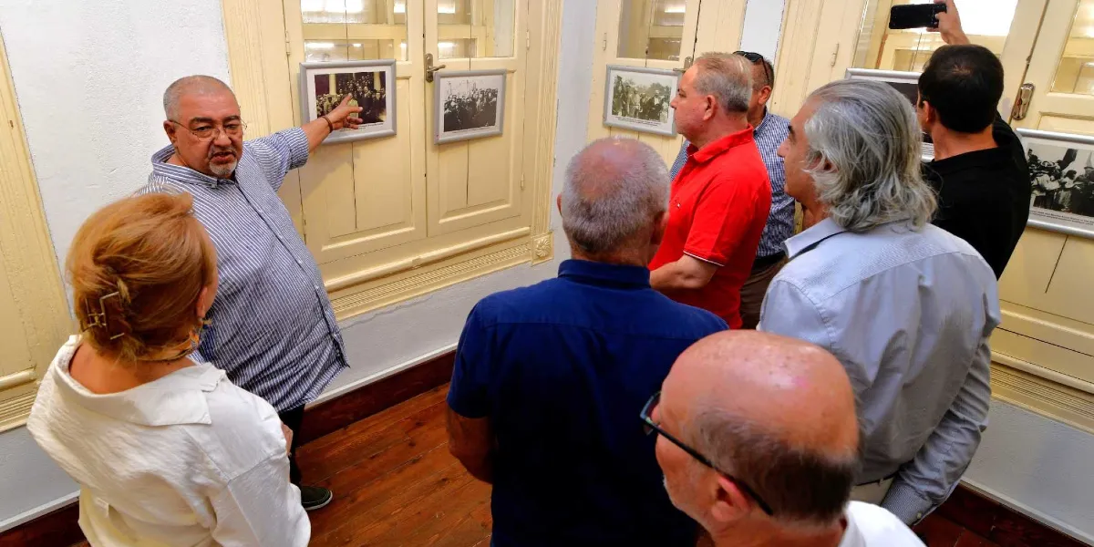 Tarihi bir binanın iç mekanında, çizgili gömlekli orta yaşlı bir adam, duvarda asılı olan siyah beyaz Atatürk fotoğraflarını işaret ederek çevresindeki bir gruba bilgi veriyor. Gruptaki diğer kişiler ilgiyle fotoğrafları incelerken, arka planda ahşap pencereler ve sergilenen diğer tarihi fotoğraflar görülüyor.
