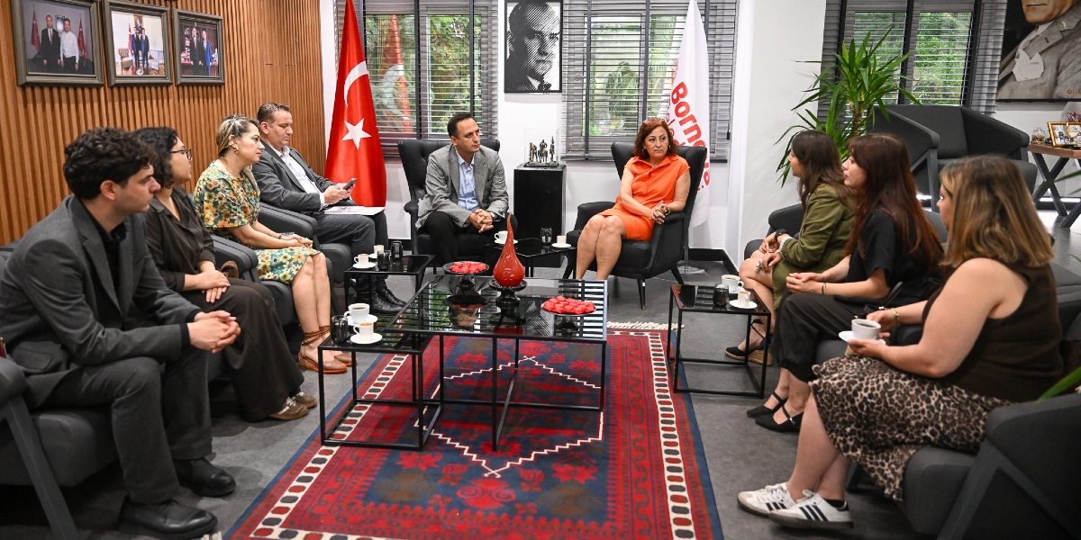 Bornova Belediyesi'nde bir toplantı odasında, karşılıklı koltuklarda oturan bir grup insan bir araya gelmiş. Odanın merkezinde kırmızı desenli bir halı ve üzerinde ikramların bulunduğu siyah bir sehpa yer alıyor. Arka planda Türk bayrağı, Bornova Belediyesi'nin flaması ve duvarda Atatürk portreleri bulunuyor. Katılımcılar, resmi ve yarı resmi kıyafetleriyle sohbet ederek toplantı gerçekleştiriyor.