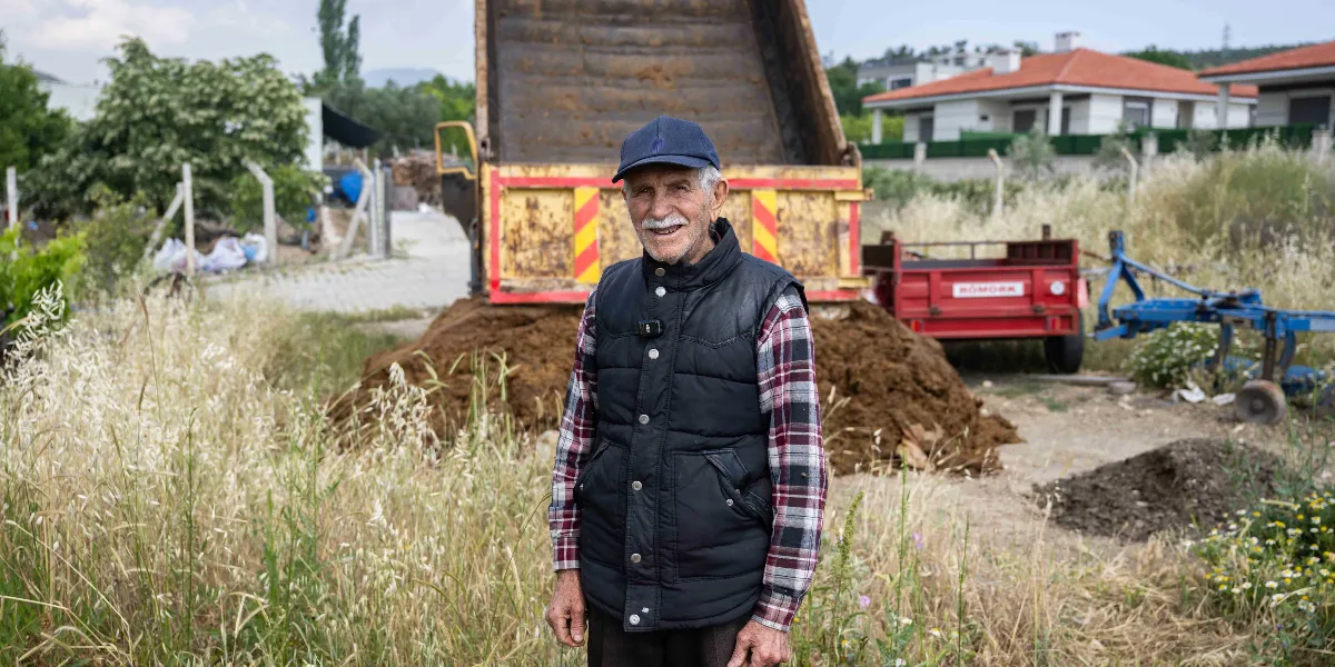 Kareli gömlek ve siyah şişme yelek giymiş, lacivert şapkalı yaşlı bir adam, arkasında büyük bir damperli kamyon ve dökülmüş gübre yığını bulunan kırsal bir alanda gülümseyerek duruyor. Arka planda kırmızı bir römork, tarım ekipmanları ve müstakil evler görünüyor.