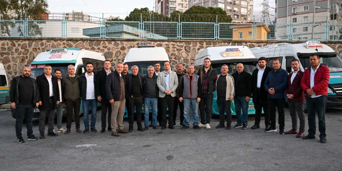 Arka planda taş duvar ve tel örgülerle çevrili bir alanda, yan yana park etmiş birkaç minibüsün önünde duran, çoğunluğu erkeklerden oluşan kalabalık bir grup. Minibüslerin üzerinde Bornova Atatürk Evka-4 güzergah bilgileri yer alıyor. Gün ışığında çekilmiş bu karede, farklı kıyafetler giymiş grup üyeleri yan yana dizilerek objektife bakıyor.