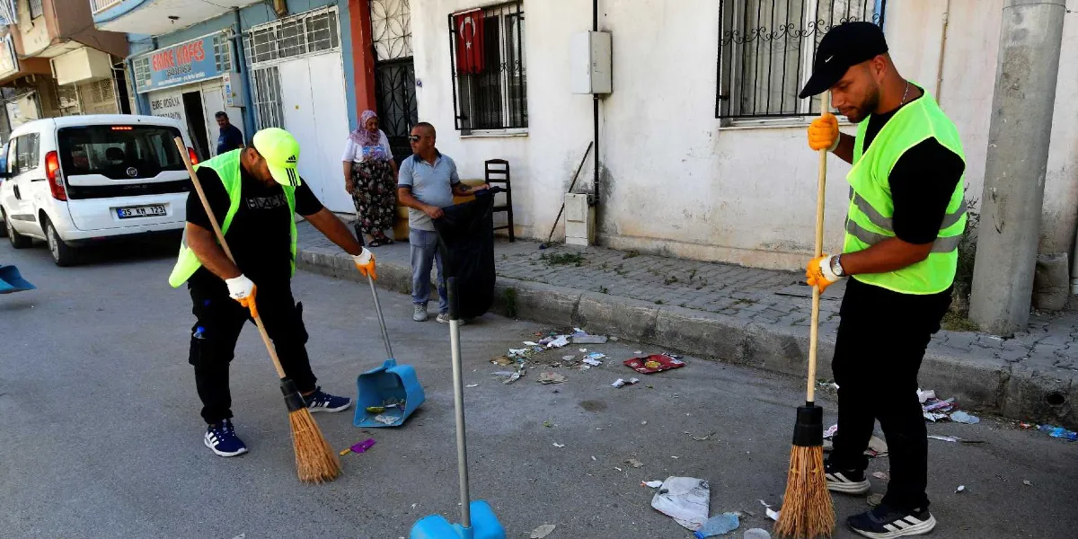Neon yeşil yelekli ve ellerinde süpürgeler bulunan iki temizlik görevlisi, Bornova'da bir sokakta yerdeki çöpleri temizliyor. Görevlilerden biri sarı şapkalı, diğeri siyah şapkalıdır. Arka planda kaldırımda duran bir kadın ve bir erkek, ellerinde çöp poşetiyle çalışmalara eşlik ediyor. Sokağın kenarında park halindeki beyaz bir araç ve duvarda asılı Türk bayrağı görülüyor.