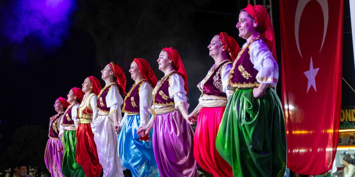 Geleneksel kıyafetler giymiş bir grup kadın, ellerini tutarak sahnede halk oyunu sergiliyor. Kadınlar; kırmızı başörtüleri, işlemeli yelekleri ve farklı renklerdeki parlak şalvarlarıyla yan yana dizilmiş durumda. Sahnenin sağ tarafında büyük bir Türk bayrağı asılı ve ortamda sahne ışıkları kullanılmış.