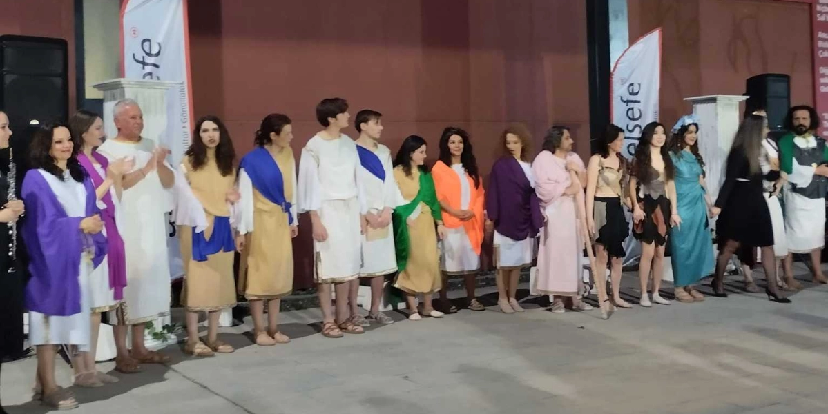 Antik Yunan dönemini anımsatan beyaz, bej, mor, turuncu ve yeşil renkli tunikler giymiş bir grup kadın ve erkek, bir sahnede yan yana dizilmiş duruyor. Bazı katılımcıların omuzlarında renkli şallar bulunurken, bir kişi elinde asaya benzer bir çubuk tutuyor. Arka planda üzerinde 