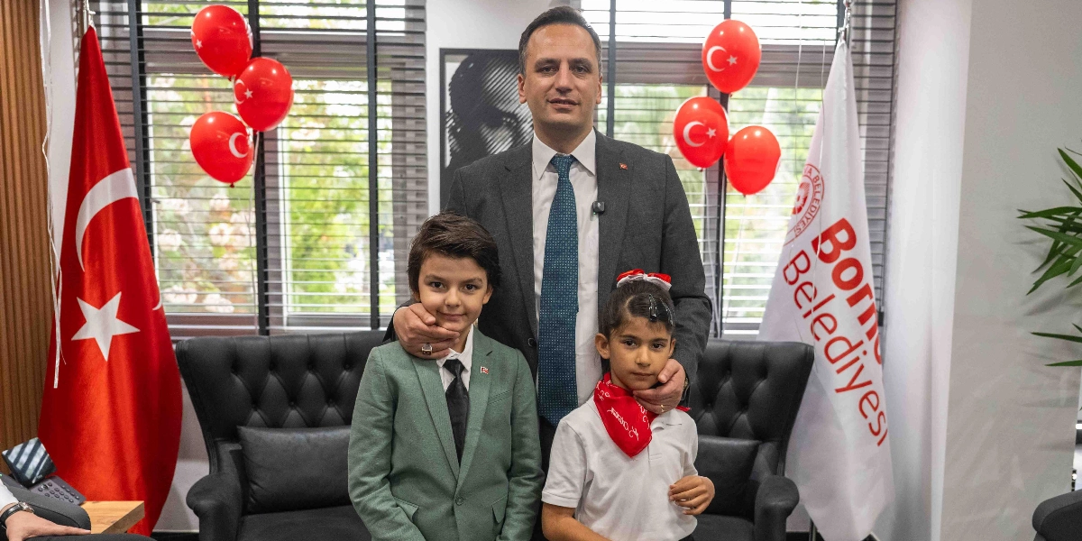 Bornova Belediye Başkanı, 23 Nisan Ulusal Egemenlik ve Çocuk Bayramı kapsamında makamında bir erkek ve bir kız çocuğuyla birlikte poz veriyor. Başkan, takım elbiseli ve kravatlı bir şekilde çocukların arkasında durarak ellerini omuzlarına koyuyor. Yanlarında Türk bayrakları ve üzerinde 