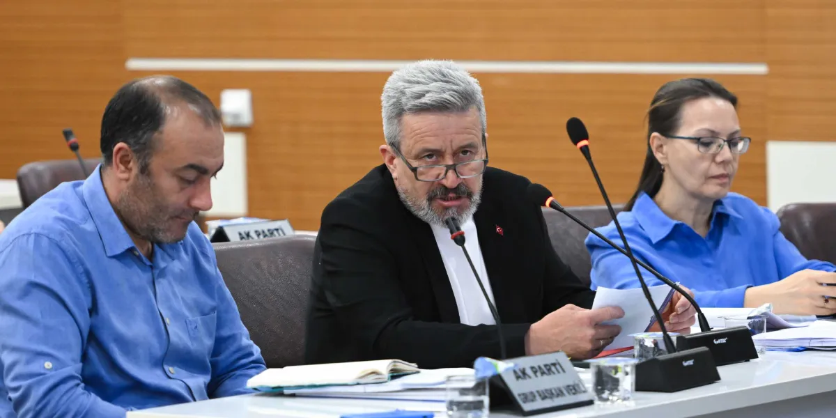 Bornova Belediye Meclisi toplantısında, ortada oturan ve konuşma yapan Meclis Başkan Vekili Tansel Ertürk, yanında oturan iki meclis üyesiyle birlikte masada yer alıyor. Ertürk siyah ceket ve gözlükleriyle mikrofon başında konuşurken, solunda mavi gömlekli bir erkek, sağında ise gözlüklü ve mavi bluzlu bir kadın oturuyor. Masanın üzerinde 