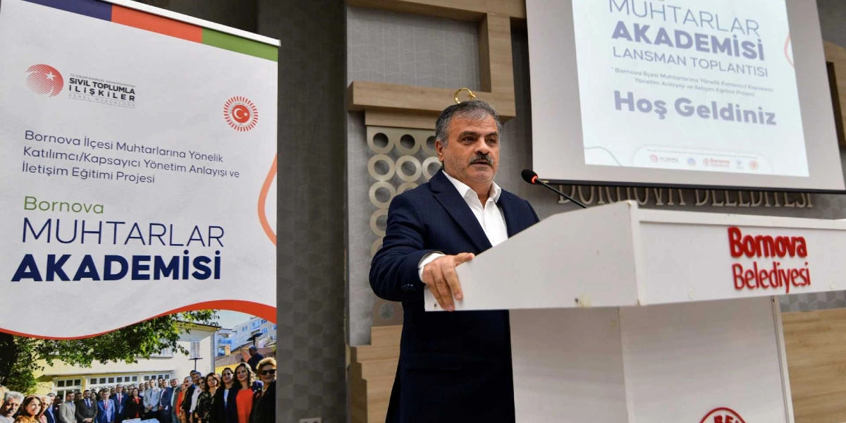 Bornova Belediyesi kürsüsünde konuşma yapan bıyıklı, orta yaşlı bir erkek. Kürsünün yanında 