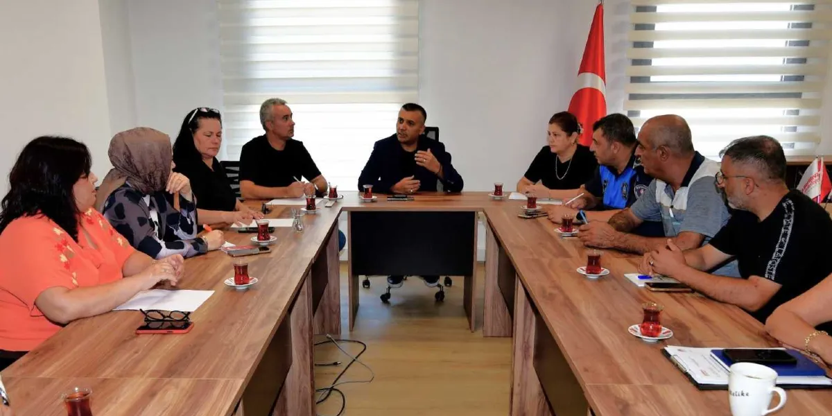 U-şeklindeki bir toplantı masasında bir araya gelmiş, aralarında Bornova Belediye Başkanı Mustafa İduğ’un da bulunduğu bir grup insan, yerel demokrasi çalışmaları kapsamında toplantı yapıyor. Katılımcılar masada otururken, önlerinde çay bardakları, not kağıtları ve kalemler bulunuyor. Arka planda Türk bayrağı ve jaluzi perdeli pencereler yer alıyor.