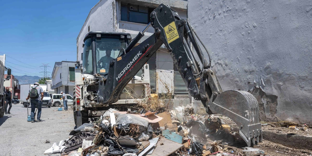 Bornova'da bir sokakta, Hidromek marka bir iş makinesi, yol kenarındaki ayakkabı atıkları ve çeşitli çöplerden oluşan yığını kepçesiyle topluyor. İş makinesinin yanında temizlik yapan görevliler bulunuyor.