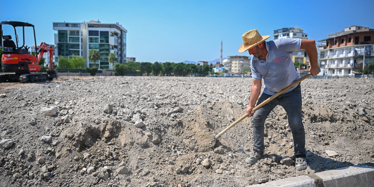 Hasır şapkalı bir adam, şehir merkezindeki boş ve taşlı bir arazide elindeki kürekle toprağı kazıyor. Arka planda modern apartmanlar, bir cami minaresi ve sol tarafta park halinde duran turuncu renkli küçük bir iş makinesi görülüyor.
