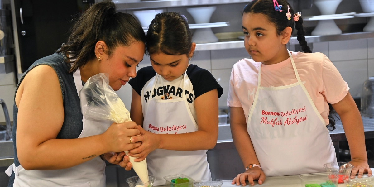 Üzerlerinde Bornova Belediyesi Mutfak Atölyesi yazılı beyaz önlükler giymiş bir eğitmen ve iki kız çocuğu, profesyonel bir mutfak tezgahında pasta süsleme çalışması yapıyor. Eğitmen, elindeki krema torbasını tutan küçük bir kızın ellerine rehberlik ederken, diğer kız çocuğu dikkatle onları izliyor.