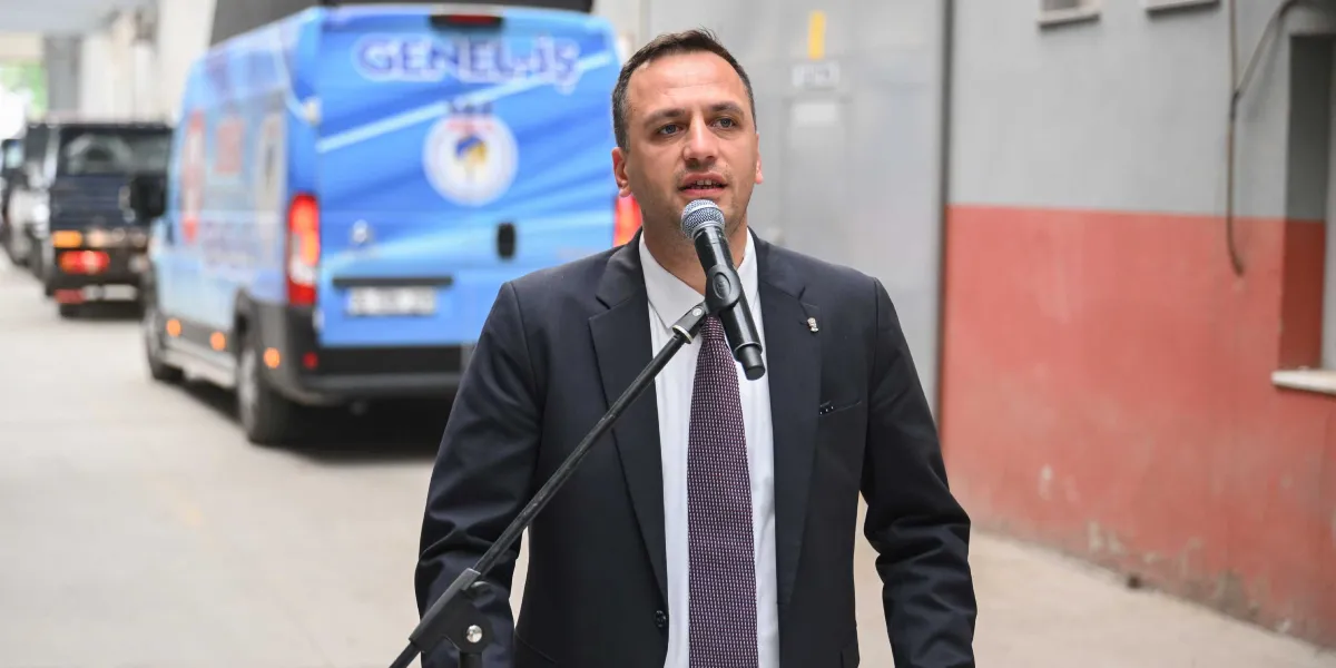 Koyu renk takım elbiseli ve kravatlı Ömer Eşki, bir mikrofon standının önünde durarak konuşma yapıyor. Arka planda üzerinde 
