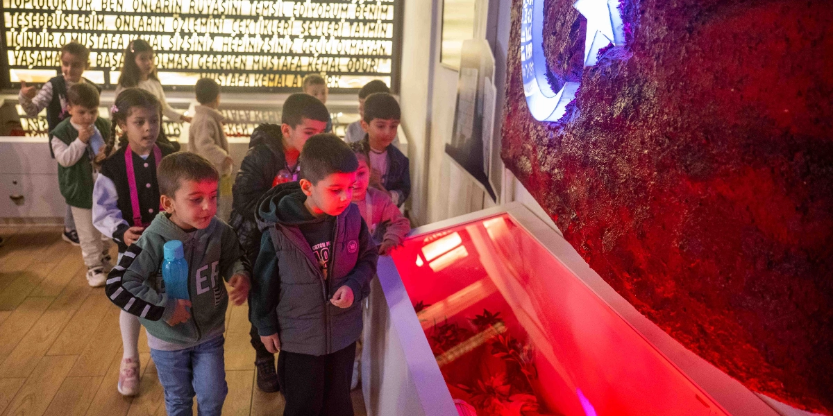 Belkahve'deki bir müze ortamında, üzerinde Mustafa Kemal Atatürk'ün sözlerinin yazılı olduğu ışıklı bir panonun önünde yürüyen ve kırmızı ışıkla aydınlatılan büyük bir Türk bayrağı rölyefini inceleyen bir grup çocuk.