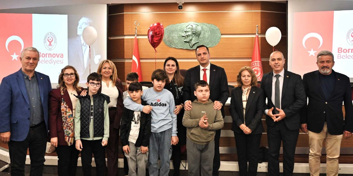 Bornova Belediyesi'nde düzenlenen 23 Nisan etkinliğinde, aralarında çocukların ve belediye yetkililerinin bulunduğu bir grup, arka planda Atatürk kabartması ve Türk bayrakları bulunan bir toplantı salonunda yan yana durarak poz veriyor. Çocuklar günlük kıyafetleriyle, yetişkinler ise resmi giysileriyle yer alıyor. Salonun iki yanında Bornova Belediyesi logolu ve Türk bayraklı ekranlar bulunuyor.