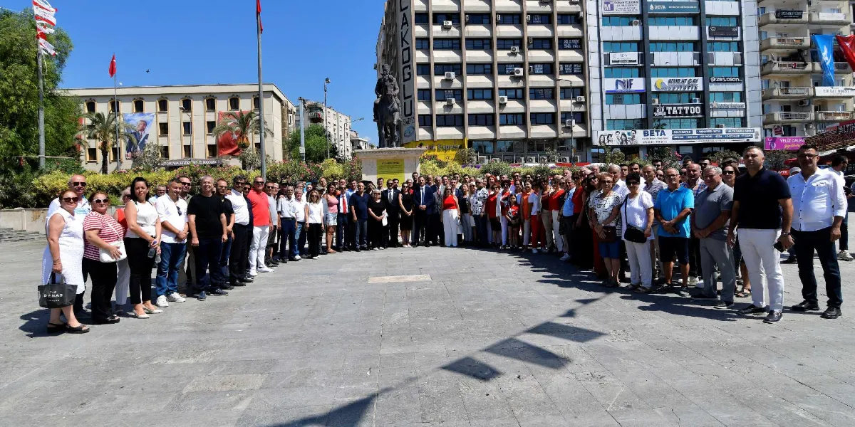 Güneşli bir günde, şehir meydanındaki Atatürk anıtının önünde bir araya gelmiş kalabalık bir grup insan yan yana durarak poz veriyor. Arka planda çok katlı binalar, Türk bayrakları ve çeşitli iş yeri tabelaları görülüyor.