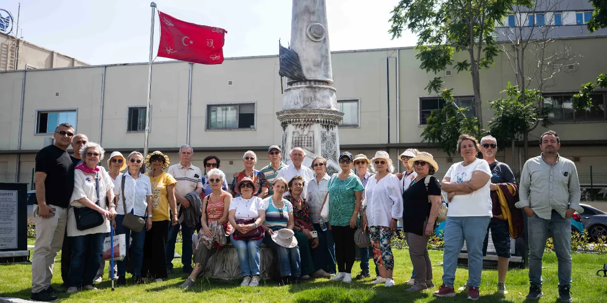 Bornova'da düzenlenen tarih ve doğa gezisine katılan, farklı yaş gruplarından oluşan kalabalık bir grup, üzerinde Osmanlıca yazılar bulunan tarihi bir anıtın önünde ve yanında yan yana durarak poz veriyor. Arka planda üzerinde ay yıldız ve Osmanlıca yazılar bulunan kırmızı bir bayrak dalgalanıyor. Grubun arkasında uzun, açık renkli bir bina ve yeşil ağaçlar yer alıyor. Katılımcıların çoğu güneş gözlüğü takıyor ve günlük kıyafetler giyiyor.