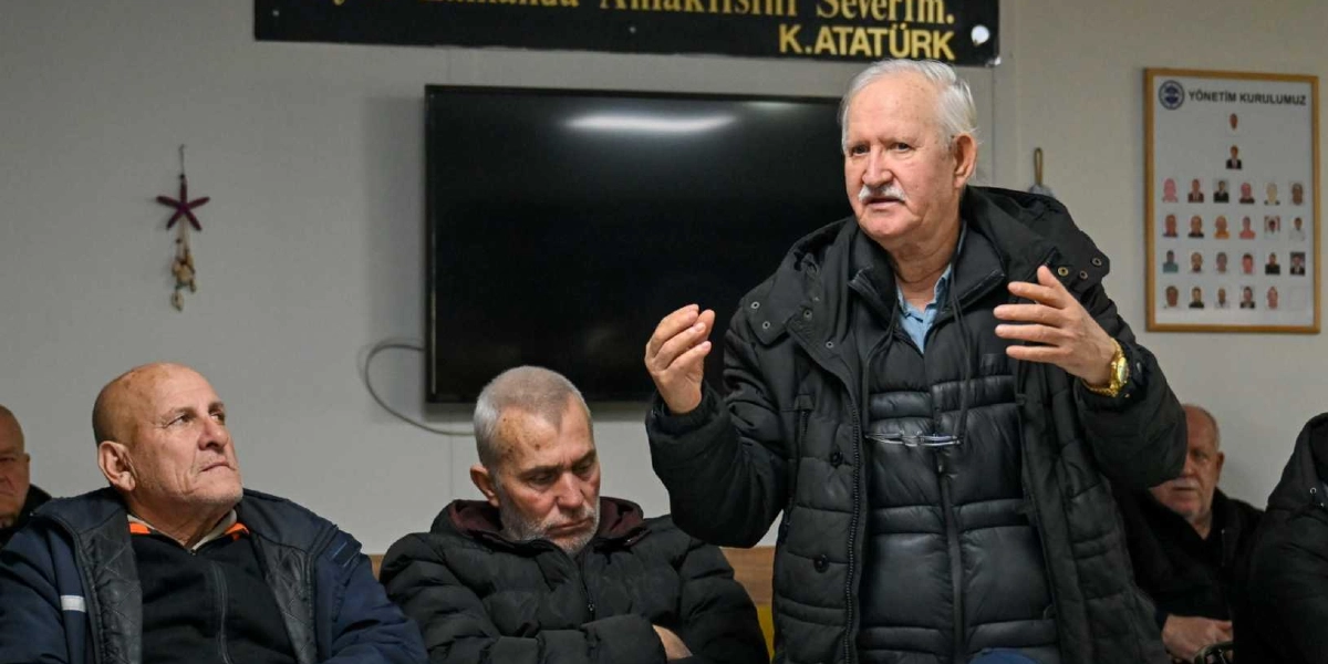 Beyaz saçlı ve bıyıklı bir adam, kapalı bir toplantı salonunda ellerini kullanarak konuşma yapıyor. Arka planda duvarda 