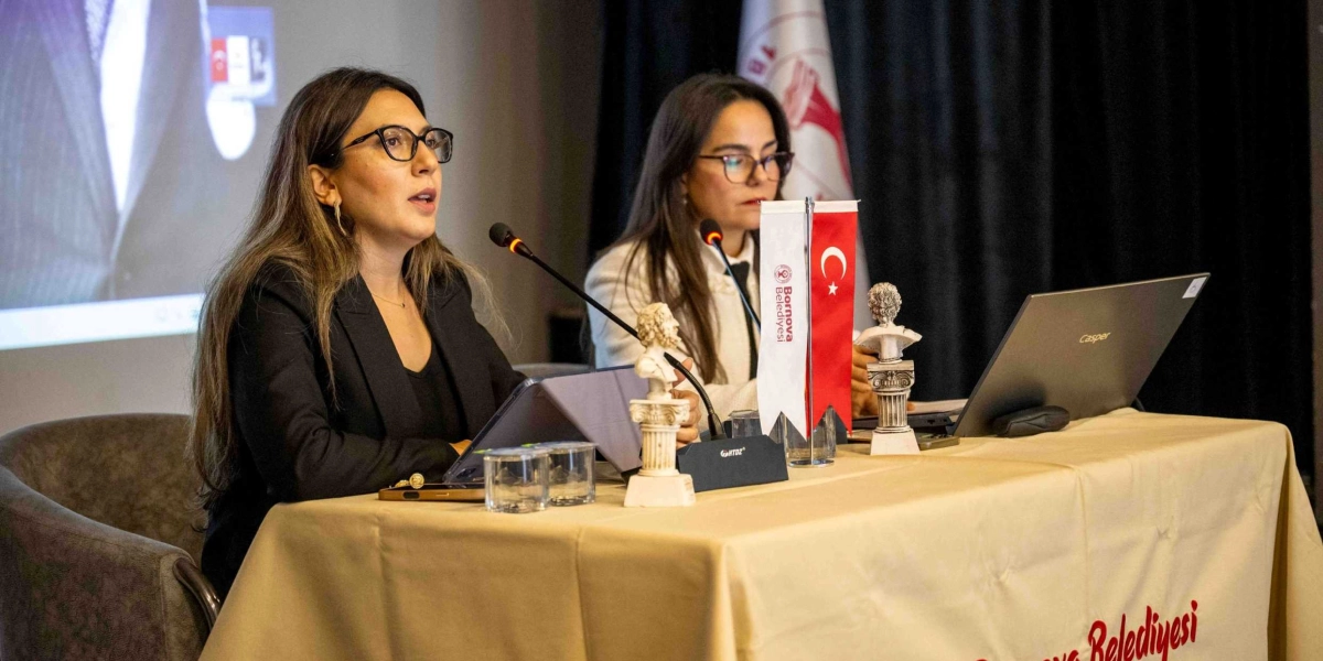 Bej renkli bir masa örtüsüyle kaplı masada yan yana oturan iki kadın konuşmacı. Soldaki kadın, siyah ceketli ve gözlüklü bir şekilde mikrofon önünde konuşma yapıyor. Sağdaki kadın ise beyaz ceketli, gözlüklü ve önünde bir Casper dizüstü bilgisayar bulunuyor. Masanın üzerinde Bornova Belediyesi logolu küçük bir Türk bayrağı, iki adet küçük büst ve mikrofonlar yer alıyor. Arka planda ise üzerinde bir figürün silüeti görünen bir projeksiyon perdesi ve siyah bir perde bulunuyor.