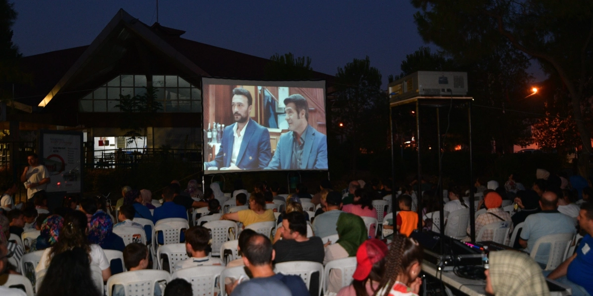 Gece vakti açık havada kurulan dev ekranda film izleyen kalabalık bir izleyici grubu. Beyaz plastik sandalyelere oturan insanlar, arkadan yansıtılan ve iki erkeğin göründüğü film sahnesine odaklanmış durumda. Arka planda büyük, çatılı bir bina ve ağaçlar yer alıyor. Sağ tarafta ise yüksek bir stand üzerinde projeksiyon cihazı ve ses sistemi bulunuyor.