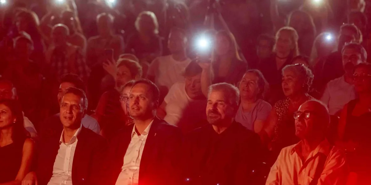 Kırmızı ışıkla aydınlatılmış bir konser veya etkinlik salonunda, ön sırada oturan takım elbiseli ve günlük kıyafetli bir grup izleyici sahneye doğru bakıyor. Arka planda ise ellerindeki telefonların ışıklarını yakarak etkinliğe eşlik eden kalabalık bir izleyici kitlesi yer alıyor.