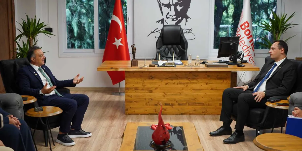 Resmi bir ofis ortamında, ahşap bir çalışma masasının karşılıklı taraflarında oturan iki takım elbiseli erkek, Atık Bank Projesi üzerine görüşme yapıyor. Arka planda duvarda Atatürk portresi, Türk bayrağı ve Bornova Belediyesi flaması yer alıyor. Masanın üzerinde küçük bir Atatürk heykeli ve isimlik bulunurken, önlerinde cam tablalı ahşap bir orta sehpa ve üzerinde kırmızı dekoratif bir obje yer alıyor.
