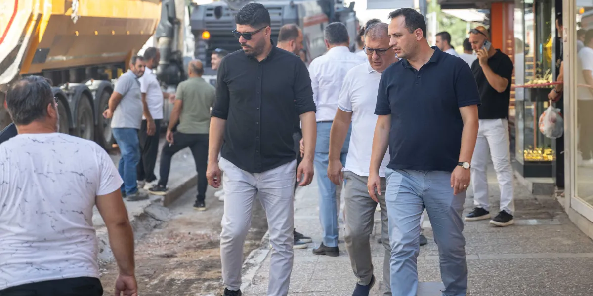 Mustafa Kemal Caddesi'ndeki yenileme çalışmaları sırasında, kaldırımda yürüyen ve incelemelerde bulunan bir grup erkek. Ön planda siyah gömlekli ve güneş gözlüklü bir kişi ile yanında lacivert polo yaka tişörtlü bir başka kişi yer alıyor. Arka planda ise iş makineleri ve caddede bulunan diğer insanlar görülüyor.