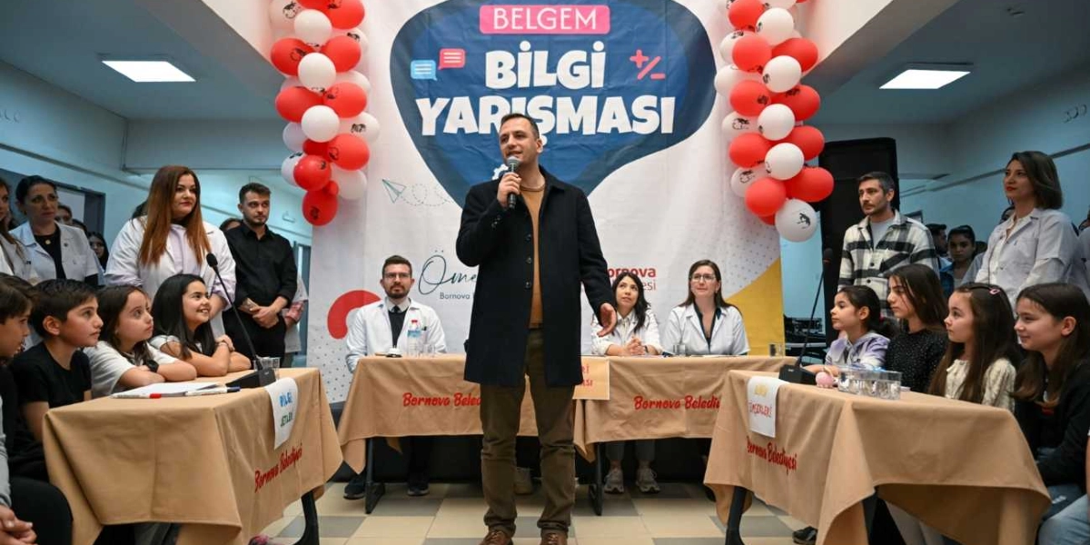 Bornova Belediyesi tarafından düzenlenen Belgem Bilgi Yarışması etkinliğinde, bir konuşmacı elinde mikrofonla ayakta dururken, önündeki masalarda oturan öğrenciler ve jüri üyeleri onu dinliyor. Arka planda 