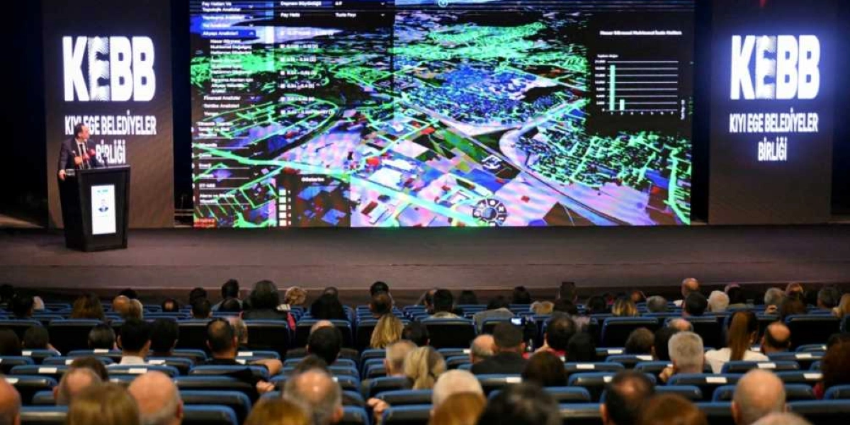 Kıyı Ege Belediyeler Birliği (KEBB) logosunun yer aldığı büyük bir sahnede, kürsüde konuşma yapan bir kişi ve arkasındaki dev ekranda afet risk analizlerini gösteren dijital bir harita yer alıyor. Harita üzerinde fay hatları, yapısal veriler ve istatistiksel grafikler bulunuyor. Salonu dolduran izleyiciler, sahnedeki sunumu takip ediyor.
